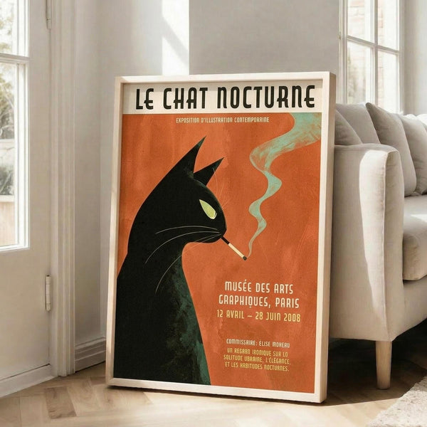 May include: Framed vintage-style poster featuring a black cat silhouette smoking a cigarette with green smoke curls against an orange background. French text reads "LE CHAT NOCTURNE" at top. The wooden-framed artwork leans against a white wall next to a beige upholstered chair in a bright, minimalist room with large windows and natural lighting.