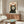 May include: Framed retro-style poster of a black and white tuxedo cat with wide yellow eyes and open mouth saying "Meow" in red text, displayed above a mid-century modern walnut credenza with brass handles. Room features white upholstered swivel chair, brass floor lamp, white table lamp, oval glass coffee table, and neutral walls with natural lighting.