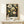 May include: Framed botanical art print with intricate illustrated flowers and seed pods in cream, yellow, and orange tones against a dark background, displayed on a light wood console table with a small wrapped gift box beside it on a neutral wall.