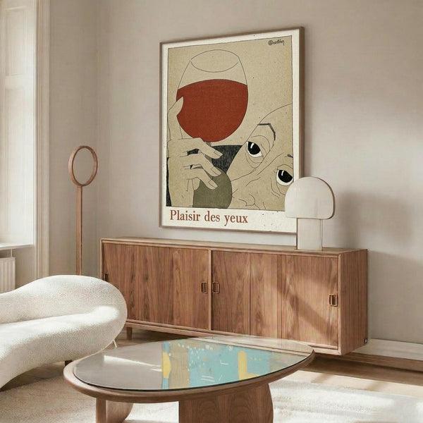 May include: Framed vintage-style art poster showing hands holding a wine glass filled with red wine, featuring stylized eyes and "Plaisir des yeux" text at bottom. Displayed above a mid-century modern walnut credenza with white mushroom table lamp, alongside a white curved chair and oval glass coffee table in a minimalist living room with cream walls.