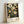 May include: Framed art print leaning against white wall showing botanical illustration with large circular flowers and detailed leaves on dark background. Light wood frame surrounds artwork featuring cream and yellow blooms with intricate centers, tall stems, and scattered dots resembling stars. Text at bottom reads "YAYOI KUSAMA" with "Modern Art 1985" and "Azure Dream Collection" details. Natural lighting creates soft shadows on wall.