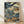 May include: Framed Japanese woodblock print artwork leaning against white wall on light wooden floor. The vintage-style print features dramatic swirling blue waves and wind patterns surrounding a small figure on a wooden bridge, with traditional architecture and trees visible in the background. Earth tones of blue, cream, and brown dominate the composition in classic ukiyo-e style, displayed in a thin wooden frame.