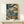 May include: Framed Japanese woodblock print showing a figure on wooden bridge amid dramatic swirling blue waves and wind patterns, displayed on light wood console table with small white wrapped gift box, against cream wall with soft natural lighting