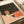 May include: Vintage-style art print showing a person with reddish hair reading in bed under a starry night sky. The illustration features warm coral and pink tones contrasting with a dark starlit background. The print appears mounted or framed, displayed alongside white paper materials and a cream ribbon on a neutral surface.