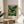 May include: Framed art poster displaying a lush green landscape with rolling hills, yellow flowering trees, and a pale sun in a golden sky. The artwork features Japanese text and "Modern Art 1950 Tokyo" at the bottom. The wooden-framed print sits on a round wooden side table next to a beige upholstered chair, with natural sunlight casting shadows across white walls and a plant visible in the corner.