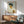 May include: Framed reproduction of an early 20th century expressionist self-portrait showing a young man with tousled dark hair and angular features, wearing a white shirt with yellow vest accent. The artwork hangs above a mid-century modern wood credenza with table lamp, next to a sage green upholstered armchair with wooden frame. Warm natural light creates geometric shadows on white wall.