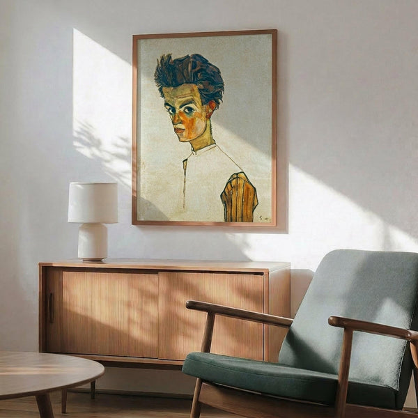 May include: Framed reproduction of an early 20th century expressionist self-portrait showing a young man with tousled dark hair and angular features, wearing a white shirt with yellow vest accent. The artwork hangs above a mid-century modern wood credenza with table lamp, next to a sage green upholstered armchair with wooden frame. Warm natural light creates geometric shadows on white wall.