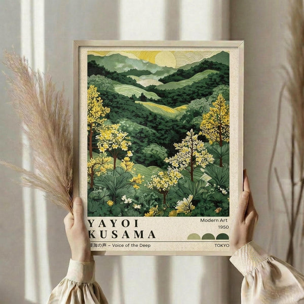 May include: Person in cream ruffled sleeve top holding framed botanical landscape artwork featuring rolling green hills, yellow flowering trees, and sunrise. Print shows layered mountain scenery in various green tones with detailed foliage in foreground. Light wood frame against neutral curtained background with dried pampas grass visible on left side.