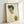 May include: Framed art print leaning against white wall showing expressionist self-portrait of young man with tousled dark hair and intense gaze. Figure wears white shirt with orange and green striped sleeve detail. Artwork features bold outlines, warm earth tones of yellow and orange flesh, set against cream background. Natural wood frame casts soft shadows on textured wall surface.
