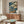 May include: Japanese woodblock print artwork displayed above walnut credenza in modern living room. Print features dramatic blue ocean waves, golden temple buildings, and figure crossing wooden bridge in traditional ukiyo-e style. Room has cream walls, white curved chair, glass coffee table, and brass floor lamp.