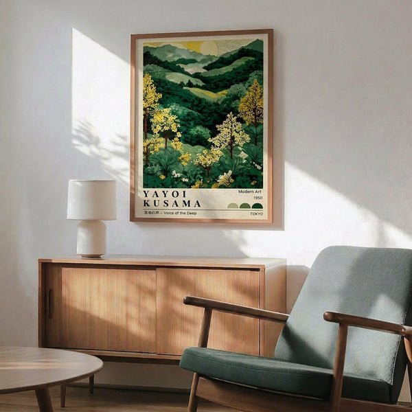 May include: Framed poster displaying a lush mountain landscape with rolling green hills, golden flowering trees in foreground, and yellow sun in sky. Print features "YAYOI KUSAMA" text and "Voice of the Deep" subtitle in exhibition poster style. Shown hanging above wooden sideboard with white ceramic lamp, next to mid-century modern chair with sage green upholstery and walnut wood arms in sunlit room with white walls.