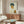 May include: Expressionist self-portrait artwork displayed above wood sideboard in modern living room setting. The painting shows a young man with dark tousled hair and angular features in white shirt, rendered in bold blues, oranges and yellows against cream background. Warm walnut furniture, white curved chair, glass coffee table, and soft natural lighting complete the contemporary interior scene.