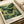 May include: Vintage-style art print featuring lush green landscape with rolling hills, flowering trees with yellow and white blossoms, and tropical foliage in Japanese artistic style. Print displayed on cream-colored paper with wide white border, text reading "YAYOI KUSAMA" and "Voice of the Deep" below artwork. Shows "Modern Art 1950 TOKYO" in corner with color palette samples. Print lies flat on neutral surface next to rolled paper tube.