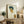 May include: Framed reproduction of an early 20th century self-portrait showing a young man with tousled dark hair and intense gaze, painted in expressive style with bold brushstrokes in blues, oranges and yellows against cream background. Wooden frame displayed on rustic sideboard with fern plant, ceramic vases, and books in neutral-toned living room setting.