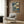 May include: Japanese woodblock print artwork displayed in modern living room setting with beige upholstered chair, gold floor lamp, and wood paneled wall. Artwork features dramatic blue swirling waters, mountains, trees, and wooden bridge in traditional ukiyo-e style with earth tones and deep blues. Print appears framed and hung above contemporary furniture on warm wood grain background.