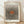 May include: Framed poster print leaning against white textured wall on light wooden floor. The artwork features radiating black polka dots emanating from a central rust-colored oval on cream background, creating an optical illusion effect. Text reads "YAYOI KUSAMA" at top and "Modern Art 1975" at bottom. Natural wood frame with subtle shadows cast on wall and floor.