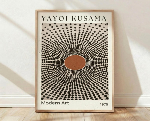 May include: Framed poster print leaning against white textured wall on light wooden floor. The artwork features radiating black polka dots emanating from a central rust-colored oval on cream background, creating an optical illusion effect. Text reads "YAYOI KUSAMA" at top and "Modern Art 1975" at bottom. Natural wood frame with subtle shadows cast on wall and floor.