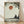 May include: Framed art print leaning against white wall on light wood floor. The artwork shows a silhouetted figure in meditation pose sitting beneath a large tree by water, with a rust-colored sun in pale sky. Rendered in muted earth tones with painterly, minimalist style. Natural wood frame, soft natural lighting creates shadows on wall.