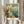 May include: Hands holding a framed vintage travel poster featuring Navy Pier with a large ferris wheel, pier extending into blue water with sailboats, and classic architecture. Warm cream and teal color palette in retro illustration style. Person wears cream sweater and holds dried pampas grass, standing near white curtained window.