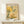 May include: Framed botanical poster with yellow and green floral design featuring dotted patterns and abstract plant motifs in Kusama's distinctive style, displayed on light wood shelf against cream wall with small wrapped gift box beside frame