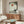 May include: Zen-inspired poster print displayed above wooden credenza in modern living room. Artwork shows silhouetted monk meditating beneath large tree with orange-red sun overhead in muted earth tones. Room features cream walls, white curved chair, oval glass coffee table with colorful reflections, and warm wood furniture with mid-century styling.