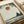 May include: Framed art print on cream paper showing a minimalist landscape with dark silhouetted figure sitting beneath tree branches, coral-red sun in pale sky, and horizontal ground lines. Print has weathered, textured edges and sits against rolled white paper on light wooden surface.