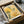 May include: Framed botanical art print in warm yellows and greens featuring stylized flowers, leaves, and dotted patterns in modern artistic style. Print shows "Modern Art" and "1999" text. Black frame sits in open cardboard shipping box with white tissue paper padding on white fabric surface.