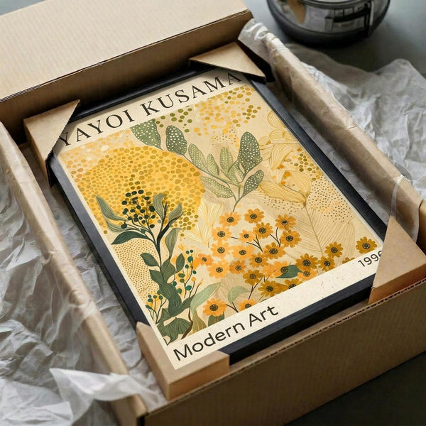 May include: Framed botanical art print in warm yellows and greens featuring stylized flowers, leaves, and dotted patterns in modern artistic style. Print shows "Modern Art" and "1999" text. Black frame sits in open cardboard shipping box with white tissue paper padding on white fabric surface.