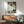 May include: Zen artwork print in wooden frame showing silhouette of meditating monk beneath tree branches with red sun above, displayed on white wall above wooden sideboard with table lamp, next to mint green upholstered armchair in modern living room setting with natural lighting and shadows