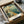 May include: Vintage-style poster artwork featuring Chicago's Navy Pier waterfront scene with Ferris wheel, sailboats on blue water, and historic pier structures in warm golden tones. Black-framed print nestled in cardboard shipping box with white tissue paper padding on neutral surface.