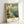 May include: Vintage-style travel poster in light wood frame leaning against white wall. Features colorful illustration of Navy Pier with large Ferris wheel, ornate domed building, sailboats on blue water, and "Welcome to Illinois" text in clouds above. Art has retro color palette with blues, creams, and warm tones in classic tourism poster aesthetic.