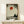 May include: Framed poster print displayed on light wood console table against white wall. Artwork shows silhouetted figure in meditation pose beneath large red sun, with dark tree branches overhead creating natural frame. Muted earth tones with weathered, vintage aesthetic. Small white gift box with twine sits on table beside frame.