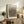 May include: Framed art print with black polka dot pattern radiating from orange center circle, displayed on rustic wooden sideboard next to potted fern, ceramic vases, and books in neutral-toned living room with beige chair and white walls