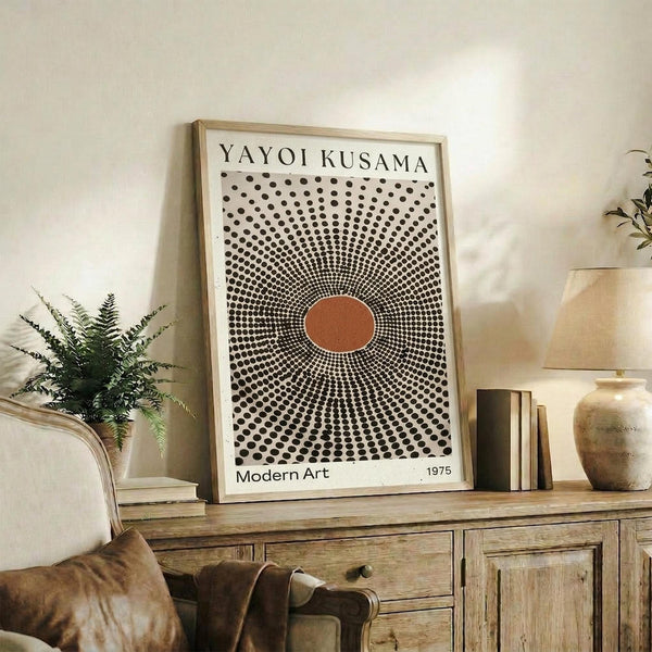 May include: Framed art print with black polka dot pattern radiating from orange center circle, displayed on rustic wooden sideboard next to potted fern, ceramic vases, and books in neutral-toned living room with beige chair and white walls