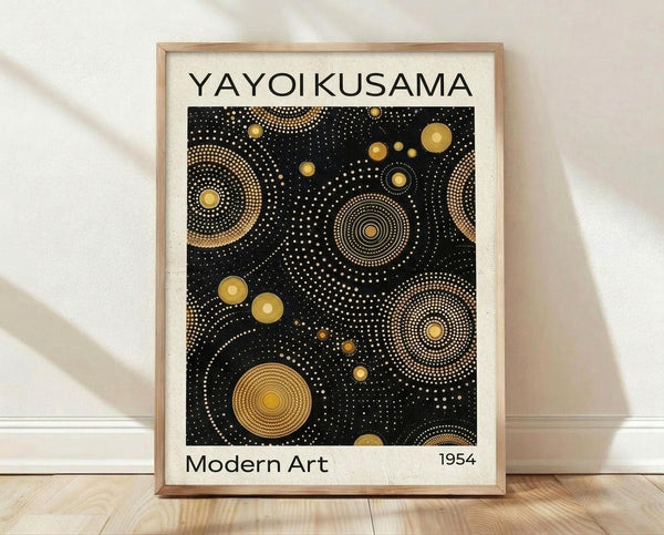 May include: Framed poster print featuring black background with golden circular patterns and dots in various sizes, creating an abstract cosmic design. The artwork shows concentric circles and dotted spirals in gold tones against dark space. Text reads "YAYOI KUSAMA" at top and "Modern Art 1954" at bottom. Light wooden frame sits against white wall on wooden floor with soft natural lighting.
