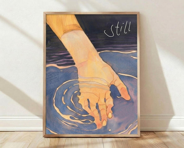 May include: Framed poster print leaning against white wall on wooden floor. Artwork shows a hand reaching into blue water creating concentric ripples, rendered in warm orange and cool blue tones with the word "still" in white text. Natural wood frame, soft window light casting shadows across the scene.