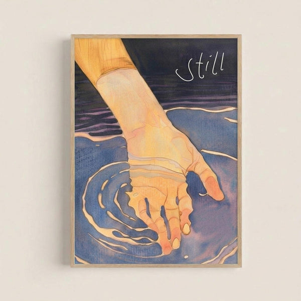 A framed art print showing a hand reaching into blue-purple water, creating ripples around the fingers. The hand is rendered in warm golden-orange tones with flowing linework, contrasting against the cool blue water. The word "still" appears in white script in the upper right corner. The artwork has a dreamy, fluid style with soft color gradients and is displayed in a light wood frame against a white wall.