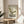 May include: Framed vintage-style design poster featuring olive green Barcelona-style chair illustration on cream background, displayed on round wooden side table next to beige upholstered chair with throw pillow. Natural sunlight streams across neutral-toned room with plant leaves visible in corner. Poster shows "1983," "Timeless Design," and "VISIONS" text in modern typography within thin wooden frame.