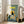 May include: Framed poster print leaning against white wall next to beige sofa in bright room with large windows. Artwork shows black cat sitting at yellow chair by breakfast table with blue checkered tablecloth, displaying plates of eggs and cookies, orange juice glasses, coffee cup, and potted plant. Yellow "CHOW!" text at top against sage green and cream background with painterly illustration style.