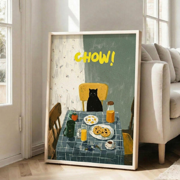 May include: Framed poster print leaning against white wall next to beige sofa in bright room with large windows. Artwork shows black cat sitting at yellow chair by breakfast table with blue checkered tablecloth, displaying plates of eggs and cookies, orange juice glasses, coffee cup, and potted plant. Yellow "CHOW!" text at top against sage green and cream background with painterly illustration style.