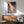 May include: Framed poster artwork showing a hand touching rippling water in warm orange and cool blue tones with "still" text, displayed above a wooden credenza with white lamp in mid-century modern living room setting with sage green armchair and natural sunlight streaming across white walls.