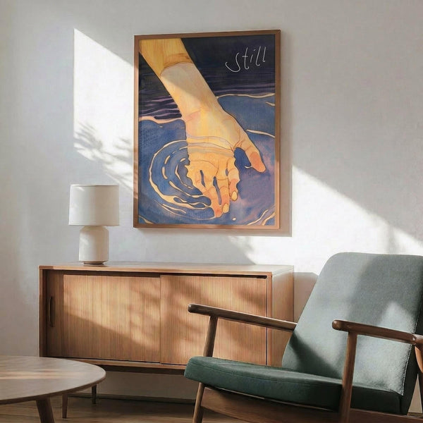 May include: Framed poster artwork showing a hand touching rippling water in warm orange and cool blue tones with "still" text, displayed above a wooden credenza with white lamp in mid-century modern living room setting with sage green armchair and natural sunlight streaming across white walls.