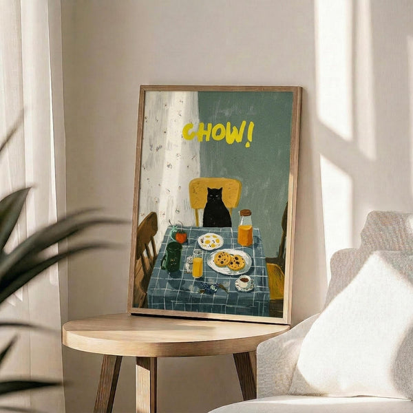 May include: Framed poster print on wooden stool next to beige armchair and plant. Artwork shows black cat sitting at breakfast table with blue checkered tablecloth, yellow chair, plates of food, coffee cup, and orange juice. Yellow "CHOW!" text at top against sage green and white background. Natural lighting creates soft shadows on white wall.