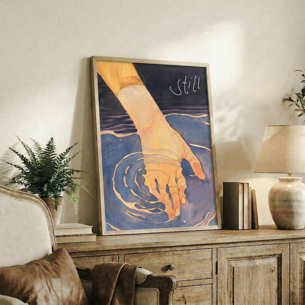 May include: Framed artwork showing a hand reaching down to touch blue water with golden ripples, creating concentric circles. The word "still" appears in white text on the dark blue background. The print sits on a rustic wooden sideboard alongside a fern plant, books, and ceramic vases in a cozy living room setting with neutral tones and natural textures.