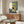 May include: Framed poster displaying a black cat sitting at breakfast table with yellow "CHOW!" text, shown in modern living room with walnut credenza, white curved chair, glass coffee table, and warm lighting. Art features checkered tablecloth, eggs, pancakes, and orange juice in folk art style with sage green and cream background.