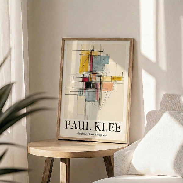 May include: Framed abstract art poster with geometric shapes in yellow, blue, red, and orange with black linear elements, displayed on a round wooden side table next to a beige upholstered chair. Natural sunlight streams across white walls, with a plant's leaves visible in the corner. The artwork features overlapping translucent rectangles and angular lines in a modernist style.