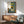May include: Framed poster showing black cat sitting at breakfast table with yellow "CHOW!" text, displayed above wooden sideboard with white lamp in modern living room setting with teal upholstered chair and natural lighting creating wall shadows