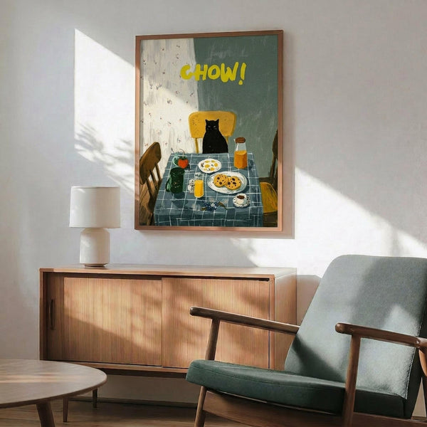 May include: Framed poster showing black cat sitting at breakfast table with yellow "CHOW!" text, displayed above wooden sideboard with white lamp in modern living room setting with teal upholstered chair and natural lighting creating wall shadows