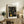 May include: Framed art print displaying black background with golden circular dot patterns and concentric rings in Yayoi Kusama's signature polka dot style, positioned on rustic wooden sideboard next to potted fern, books, and ceramic vase in warm-toned living room setting with beige upholstered furniture