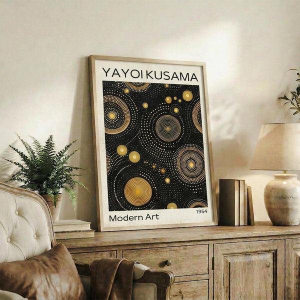 May include: Framed art print displaying black background with golden circular dot patterns and concentric rings in Yayoi Kusama's signature polka dot style, positioned on rustic wooden sideboard next to potted fern, books, and ceramic vase in warm-toned living room setting with beige upholstered furniture